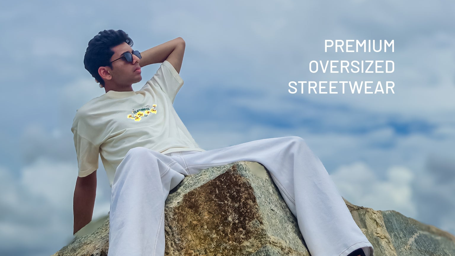 male model wearing a cream oversized t-shirt with Valour Clothing Co. logo, sitting outdoors; representing premium streetwear and luxury fit hero banner
