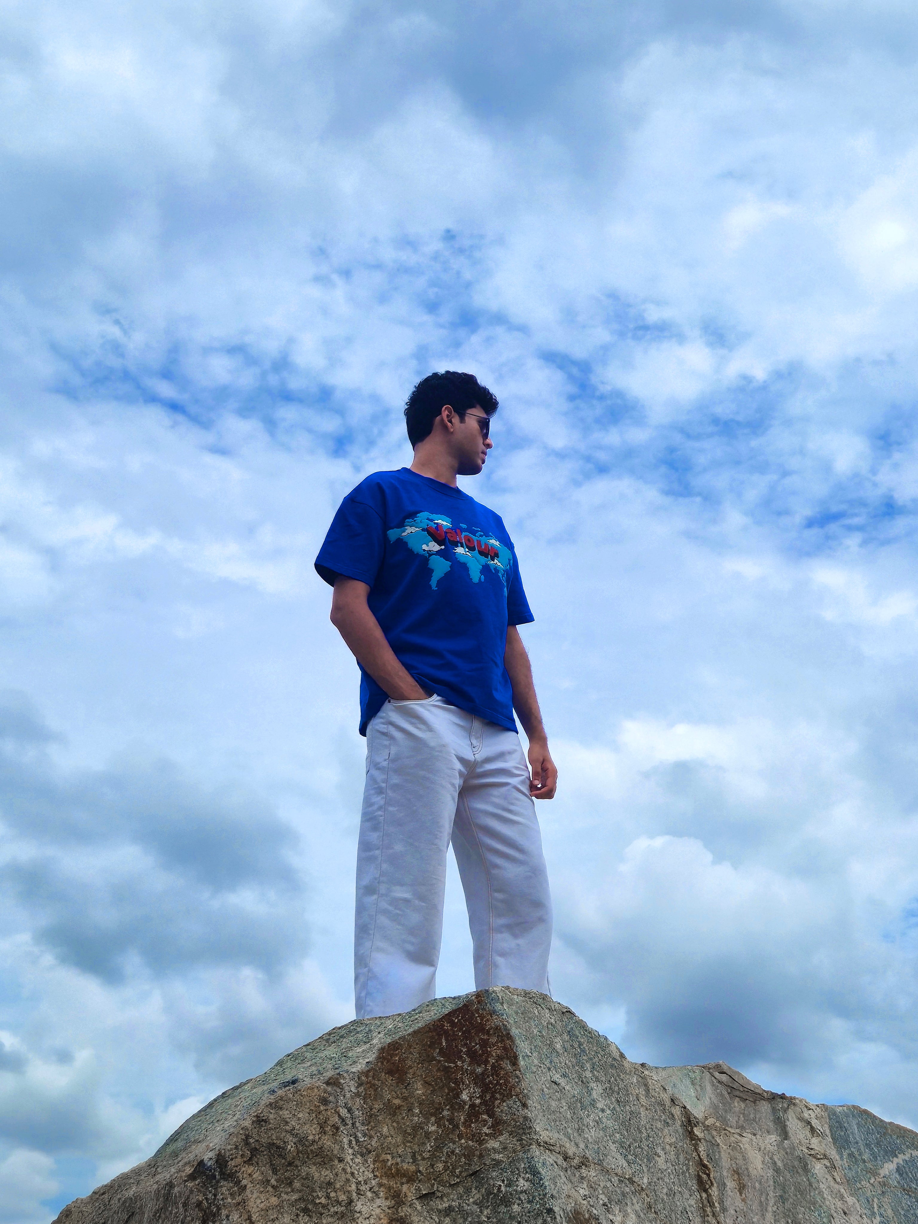 male model wearing a vibrant blue oversized t-shirt with world map graphic and white wide-leg jeans, standing on a rock under a cloudy sky
