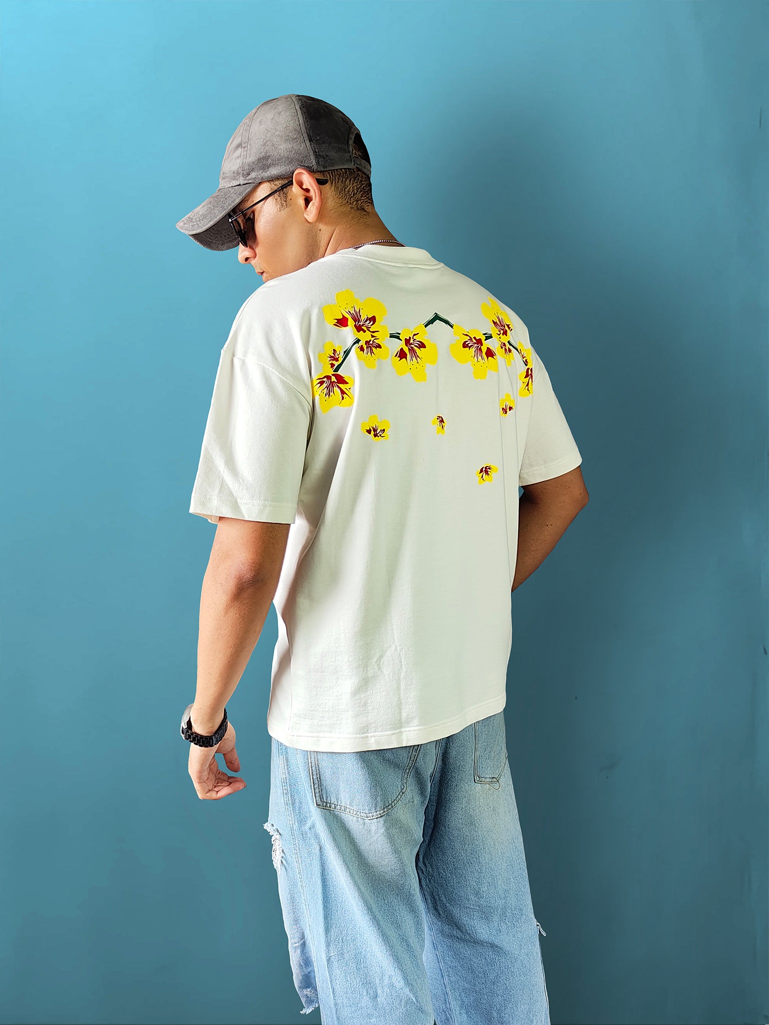 male model from behind wearing a cream oversized t-shirt with a large yellow and red floral graphic across the upper back, styled with baggy denim shorts