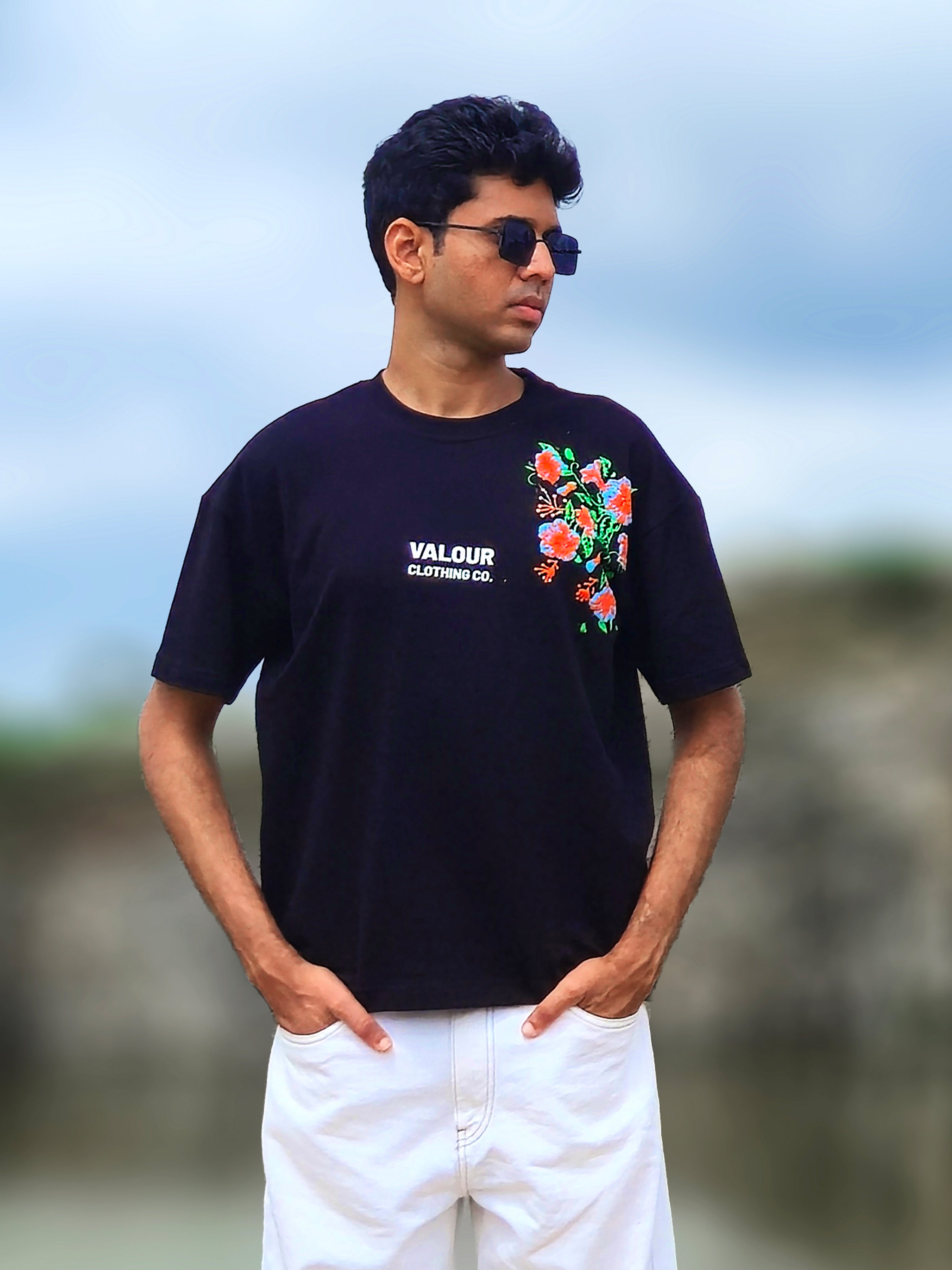 male model wearing a black oversized t-shirt with a side panel red and green floral graphic and white Valour Clothing Co. text, styled with white jeans