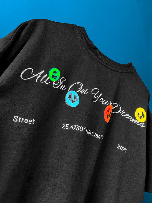 black oversized t-shirt with white script "All In On Your Dreams" and colorful graphic Valour logo, including latitude longitude coordinates and street style text